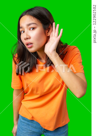 Young woman in orange shirt curiously listening against a bright green backdrop capturing attention 122902582