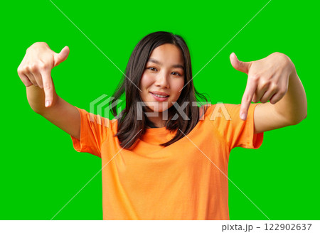 Young woman in orange shirt gestures playfully against bright green backdrop while smiling and having fun Young woman in orange shirt gestures playfully against bright green backdrop while smiling and having fun 122902637
