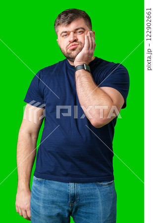 Young man with a contemplative expression leaning against green backdrop while resting his head on his hand 122902661