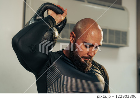 Bald man lifting kettlebell with focused expression in a gym setting during workout session Bald man lifting kettlebell with focused expression in a gym setting during workout session 122902806