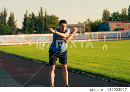 Young man stretches on the athletic track in a sunny stadium during evening practice session Young man stretches on the athletic track in a sunny stadium during evening practice session 122903067