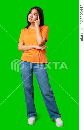 Young woman in orange shirt standing with hand on chin against bright green background 122903449