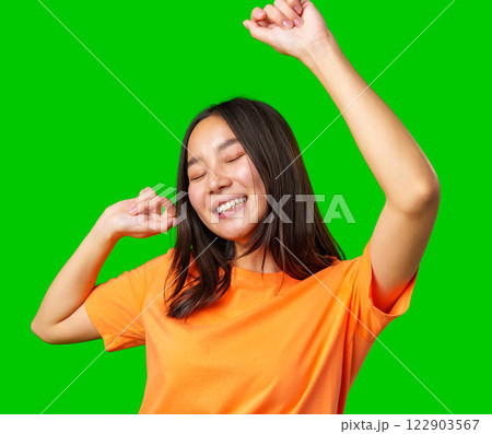 Joyful young woman dancing happily in a bright orange shirt against a green background 122903567