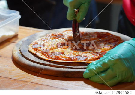 A person wearing green gloves is cutting thin-crust, small-size pizzas on a wooden tray with a pizza cutter. 122904580
