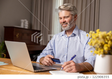 Gray-haired mature man sitting at the laptop and making notes while working Gray-haired mature man sitting at the laptop and making notes while working 122906355