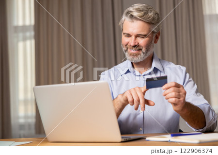 Gray-haired mid aged bearded man sitting at the table and making online payments Gray-haired mid aged bearded man sitting at the table and making online payments 122906374