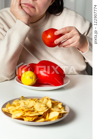 Vertical shot of Woman being tired of dieting. Boring healthy food. Overweight female choosing between vegetables and fried chips. Weight loss and diet concept. Vertical shot of Woman being tired of dieting. Boring healthy food. Overweight female choosing between vegetables and fried chips. Weight loss and diet concept. 122907774