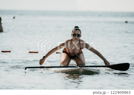 Sea woman sup. Silhouette of happy middle aged tanned woman in rainbow bikini, surfing on SUP board, confident paddling through water surface. Idyllic sunset. Active lifestyle at sea or river. 122909963