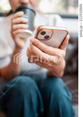 A person in casual attire holds a smartphone while sipping coffee, capturing a moment of relaxation and connectivity. A person in casual attire holds a smartphone while sipping coffee, capturing a moment of relaxation and connectivity. 122910889