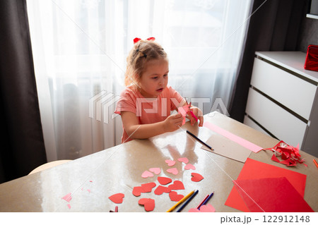 Top view of cute little Caucasian girl is sitting at a table and cutting out paper hearts. Child learns how to make crafts for Mother's Day and Valentine's Day 122912148