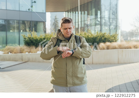 Portrait of serious 30s man with backpack pointing at his smart watch on the street in the city. Young upset casual man waiting for someone outdoors 122912275