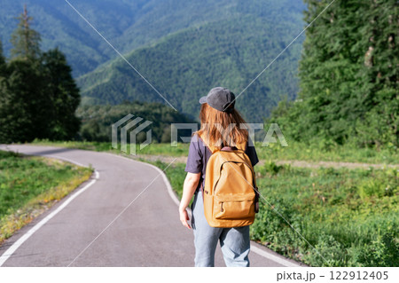 female hiker in casual clothing walks along remote asphalt road with stunning view of green mountains, Woman with backpack Summer adventure and outdoor lifestyle , travel tourism concept , copy space 122912405