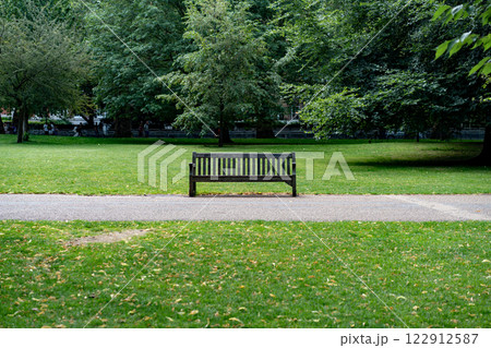 A dark wooden bench sits in St James Park along a pathway surrounded by green grass and large trees. This tranquil image is perfect for promoting urban parks and outdoor relaxation spaces. A dark wooden bench sits in St James Park along a pathway surrounded by green grass and large trees. This tranquil image is perfect for promoting urban parks and outdoor relaxation spaces. 122912587