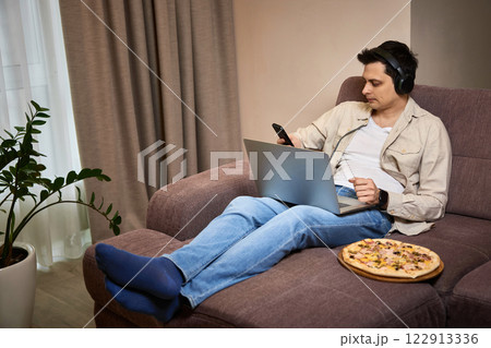 Young man sitting on sofa and eating pizza while working on laptop 122913336