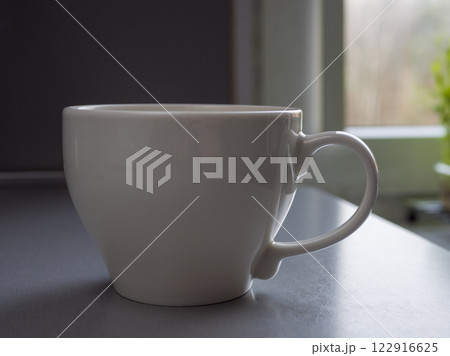 Porcelain white coffee mug steaming near sunlit window, casting warm glow across wooden surface with soft morning illumination Porcelain white coffee mug steaming near sunlit window, casting warm glow across wooden surface with soft morning illumination 122916625