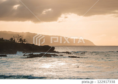 A serene coastal scene at sunset on Oahu, Hawaii, featuring gentle waves, a lone swimmer, silhouetted palm trees, and distant mountain ridges. 122917338