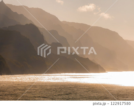 The Napali Coast on Kauai Island features jagged cliffs in golden light, silhouetted against the sky with the ocean reflecting the warm hues below. The Napali Coast on Kauai Island features jagged cliffs in golden light, silhouetted against the sky with the ocean reflecting the warm hues below. 122917580