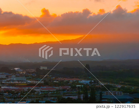 A vibrant sunset illuminates a small town on Kauai island, Hawaii, with orange and yellow skies, lush greenery, and distant mountain silhouettes. 122917635