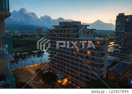 A cityscape of Honolulu at dusk featuring a lit multi story building, a canal lined with palm trees, and mountains silhouetted against the sky. A cityscape of Honolulu at dusk featuring a lit multi story building, a canal lined with palm trees, and mountains silhouetted against the sky. 122917644