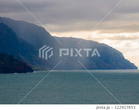 Towering cliffs of the Napali Coast meet the calm Pacific Ocean under an overcast sky, with sunlight highlighting the lush, rugged terrain. 122917653