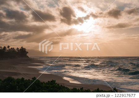 A serene Hawaiian beach at sunset with golden light over ocean waves, scattered clouds, lush greenery, palm trees, and distant hills. A serene Hawaiian beach at sunset with golden light over ocean waves, scattered clouds, lush greenery, palm trees, and distant hills. 122917677
