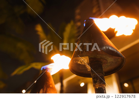 Close up of lit tiki torches with flames, set against a tropical nighttime backdrop. Palm fronds and a faint building outline are visible in the background. Close up of lit tiki torches with flames, set against a tropical nighttime backdrop. Palm fronds and a faint building outline are visible in the background. 122917678