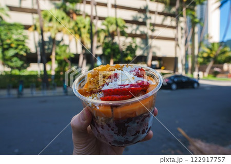 A colorful acai bowl with strawberries, mango, coconut, and granola is held against a modern building, palm trees, and a street in Honolulu. 122917757
