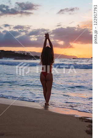 A woman stands on a sandy beach at sunset with arms raised, facing the ocean. Vibrant orange and pink hues reflect on the water, with cliffs in the distance. A woman stands on a sandy beach at sunset with arms raised, facing the ocean. Vibrant orange and pink hues reflect on the water, with cliffs in the distance. 122917761