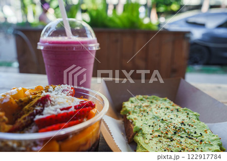 A colorful acai bowl with fruit, avocado toast, and a pink smoothie on a table. Blurred greenery and a parked car suggest a tropical setting. A colorful acai bowl with fruit, avocado toast, and a pink smoothie on a table. Blurred greenery and a parked car suggest a tropical setting. 122917784