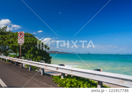 A tropical coastal road with a white guardrail runs beside a turquoise ocean under a clear sky, with lush greenery and islands on the horizon. 122917786