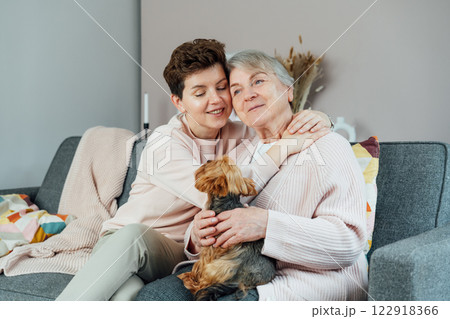Smiling senior 70s woman with daughter, Yorkshire terriers dog pet hugging on sofa. Family weekend together. Parenting love, care, generations unity concept. Spending quality time in retiring house. Smiling senior 70s woman with daughter, Yorkshire terriers dog pet hugging on sofa. Family weekend together. Parenting love, care, generations unity concept. Spending quality time in retiring house. 122918366