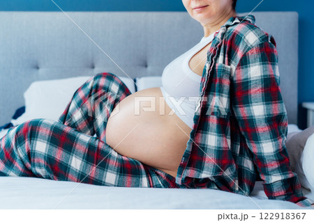 Pregnant woman sitting on bed, touching her belly, showcasing joy and anticipation. Expecting mother relaxing in pajama during last trimester of pregnancy. Motherhood, preparation and expectation 122918367