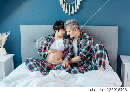 Mixed race couple are waiting for a baby. Husband and his pregnant wife in cozy pajamas relaxing on the bed at modern home bedroom. Romantic moments for pregnant couple, warm atmosphere of love. 122918368