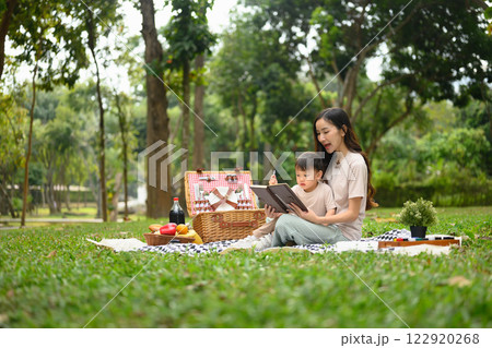 Loving mother sitting with her little son on a picnic blanket in a green park and reading book 122920268