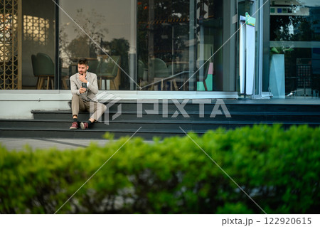 Young professional in suit checking smartphone sitting outdoors at office building Young professional in suit checking smartphone sitting outdoors at office building 122920615