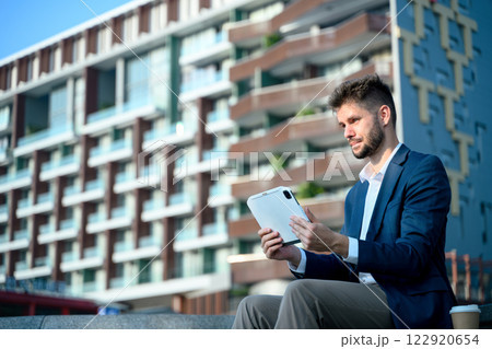 Young businessman analyzing work on his digital tablet while sitting outside a modern office building 122920654