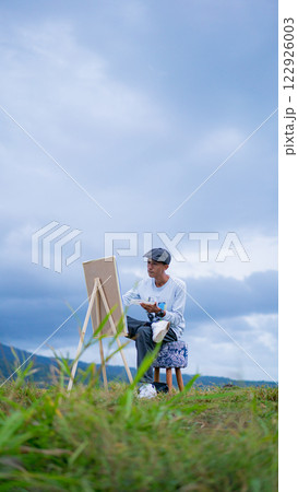 Artist painting landscape en plein air on a hilltop cloudy sky and green grass, 122926003
