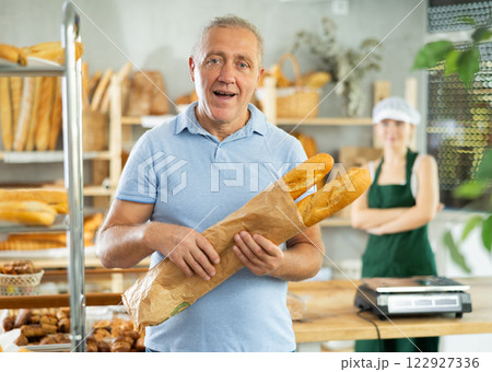Portrait of smiling male customer holding paper bag of fresh baguettes in bakery interior Portrait of smiling male customer holding paper bag of fresh baguettes in bakery interior 122927336
