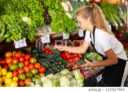 Focused girl who works part-time as a trainee saleswoman puts cucumbers on showcase Focused girl who works part-time as a trainee saleswoman puts cucumbers on showcase 122927464