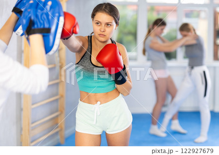 Boxing training - teen girl wearing boxing gloves exercise and punch to pads for boxing in gym Boxing training - teen girl wearing boxing gloves exercise and punch to pads for boxing in gym 122927679