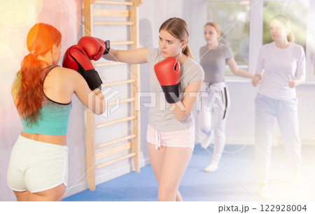 Two teenage girls in sportswear are engaged in boxing sparring in the gym under guidance of trainer in gym Two teenage girls in sportswear are engaged in boxing sparring in the gym under guidance of trainer in gym 122928004