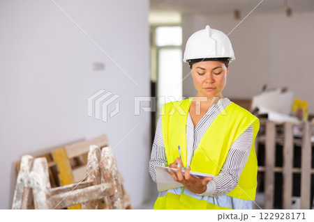 Woman foreman in a protective helmet and yellow vest checks the completed construction work on drawing Woman foreman in a protective helmet and yellow vest checks the completed construction work on drawing 122928171