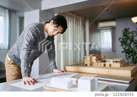 建築を設計する事務所の建築模型の置かれたデスクで建物の図面を製図する設計士の男性 122928333