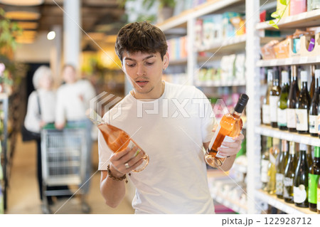 Young male customer in supermarket examines bottles of wine, chooses alcoholic drink. Young male customer in supermarket examines bottles of wine, chooses alcoholic drink. 122928712