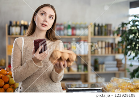 Young beautiful woman shopper in casual clothes chooses package of eggs and scanning QR with smartphone to get information about 122929109