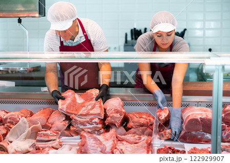 Workers of butchery shop arranging raw meat cuts in glass showcase 122929907