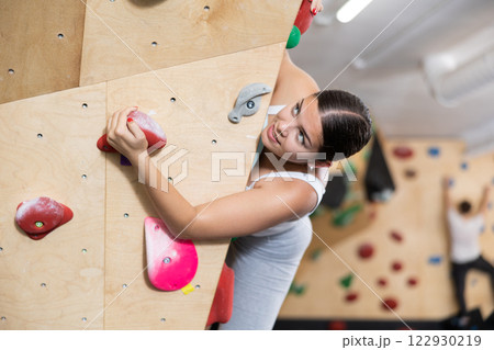 Europian girl client of gym trains doing exercises on artificial wall for climbing, top view Europian girl client of gym trains doing exercises on artificial wall for climbing, top view 122930219