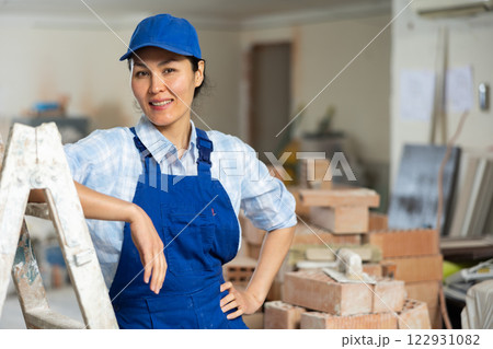 Portrait of positive builder woman in blue overalls next to stepladder 122931082
