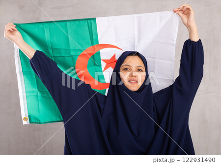 Young Muslim woman in blue hijab holds unfurled flag of Algeria. 122931490