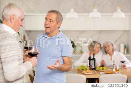 Elderly men have withdrawn from their female companions, stand with glass of wine in hands, chatting Elderly men have withdrawn from their female companions, stand with glass of wine in hands, chatting 122931560